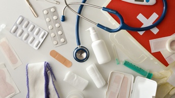 Verschiedene Objekte für Erste Hilfe und medizinische Versorgung nebeneinanderliegend, zum Beispiel ein Verband, Tablettenblister, Pflaster, ein Stethoskop, Fieberthermometer, Verbandsschere und dergleichen.