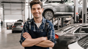 Porträt einer lächelnden Person in einem Overall und mit verschränkten Armen, die in einer großen Halle steht, in der mehrere Fahrzeuge stehen