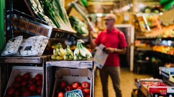 Im Vordergrund sind in und auf aufgestellten Kisten Tomaten, Bananen und Pilze. Im Hintergrund ist verschwommen eine Person mit Brille, die in der Gemüseabteilung eines Supermarktes steht. In der rechten Hälfte ist Obst