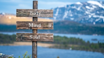Holzwegtafel mit der Beschriftung Body, Mind, Soul, im Hintergrund verschwommen Seenlandschaft mit beschneiter Bergkette