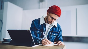 Eine Person mit roter Haube, Brille und Jeansjacke sitzt an einem Tisch und schreibt an einem Tisch neben einem Tablet in ein Notizbuch