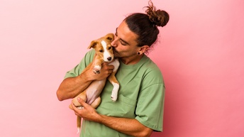Person mit dunklen, geschlossenen Haaren, Bart und T-Shirt kuschelt mit einem kleinen Welpen in der Hand während vor einem rosa Hintergrund gestanden wird