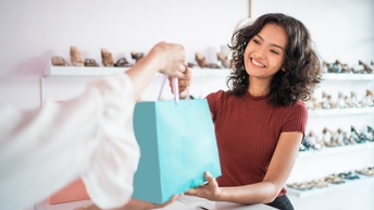 Lächelnde Person bekommt von anderer Person blaue Papiertasche über Tresen ausgehändigt, im Hintergrund verschwommen Regale mit Schuhen