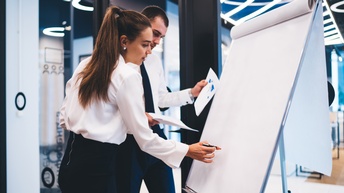 Zwei Personen in Bürokleidung stehen nebeneinander an einem Whiteboard. Die rechte Person notiert etwas mit einem Stift darauf. Die linke Person hält verschiedene Zettel