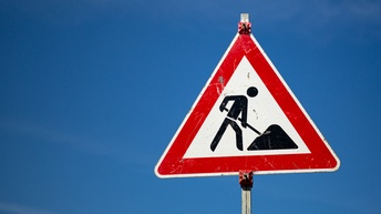 Ein dreieckiges Schild, auf dem die Silhouette einer Person eine Schaufel hält und sich nach vorne beugt. Vor ihr liegt ein Haufen. Dahinter ist der blaue Himmel