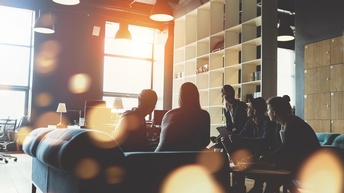 Gruppe von Personen sitzt rund um einen Tisch in einem Büro und hält eine Besprechung. Die Sonne scheint so durch das Fenster, dass nur die Silhouetten der Personen zu erkennen sind.