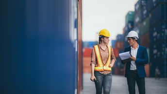 Zwei Personen mit Schutzhelm stehen auf einem Platz mit vielen Containern, eine Person trägt eine Schutzweste und hält ein Walkie-Talkie in Händen, die andere Person in Businesskleidung hält ein Klemmbrett in Händen