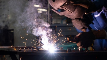 Eine Person mit Schweißhelm und Brille sowie dicken Arbeitshandschuhen beugt sich über eine Arbeitsplatte, um mit einem Schweißgerät etwas zu bearbeiten. Man sieht Funken und einen sehr hellen Punkt in der Bildmitte, wo geschweißt wird