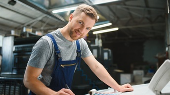 Bärtige sanft lächelnde Person in Arbeitslatzhose steht mit Stift an Tisch, Farbfächer vor sich liegend