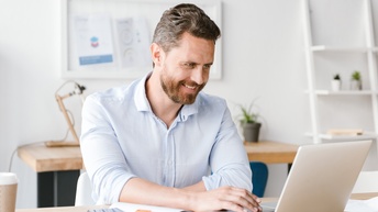 Person mit Bart und hellblauem Hemd sitzt freudig an einem Laptop während daneben ein aufgeschlagenes Notizbuch mit einem Stift und ein Smartphone liegt, im Hintergrund steht ein weiterer Schreibtisch, ein Regal sowie hängt eine Pinnwand an der Wand