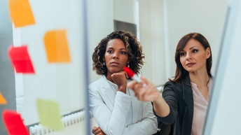 Zwei Personen stehen neben einer Glasscheibe, auf der mehrere, bunte Post-its kleben. Die rechte Personen deutet mit einem Stift auf die Post-its. Beide blicken darauf. Im Hintergrund sieht man eine Heizung und ein Fenster mit Jalousien