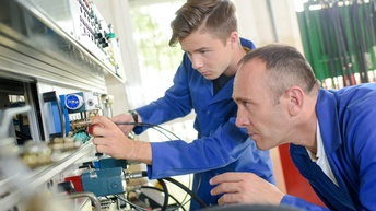 Seitliche Aufnahme zweier Personen in einem blauen Overall, die neben verschiedenen elektronischen Geräten mit Kabeln und Knöpfen. Die rechte Person hat die Hände auf einem Knopf