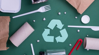 Mittig Recyclingzeichen − drei im abgerundeten Dreieck verlaufende Pfeile − umgeben von verschiedenen Verpackungsmaterialen wie Tüten, Dosen, WC-Papierrollen, Strohhalmen, Deckeln, Tüten und Einwegbesteck aus Papier und Plastik auf grünem Hintergrund