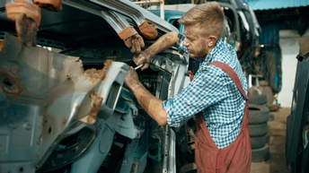 Person in Arbeitslatzhose schraubt an verschrottetem Fahrzeug
