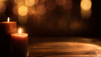 In der linken Bildhälfte stehen auf einer hölzernen Unterfläche zwei brennende Kerzen. Im Hintergrund sind hell leuchtende Punkte