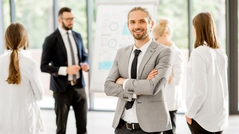 Portrait einer Person mit geschlossenen Haaren und Bart in Businesskleidung, im Hintergrund stehen weitere Personen rund um ein Flipchart
