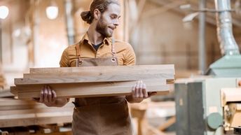 Lächelnde Person mit Bart in brauner Schürze blickt zur Seite und hält aufeinandergestapelte Holzbretter in Händen, im Hintergrund verschwommen Werkhalle