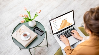 Person sitzt auf einem Sofa und blickt auf ihr Notebook mit einer Tabellen-Grafik auf ihrem Schoß, daneben steht ein Beistelltisch mit einer Blumenvase, einem Work at Home Buch sowie einem Smartphone