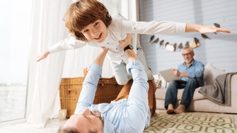 Schulkind mit braunen Haaren macht eine Fluggeste mit den Händen, während es von einer anderen Person am Boden gehalten wird, im Hintergrund sitzt eine Person mittleren Alters auf einem Sofa in einem Wohnzimmer