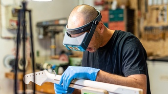 Eine Person mit Handschuhen und Schutzbrille hält einen hellen Holzgriff einer Gitarre und bearbeitet dieses mit einem Werkzeug