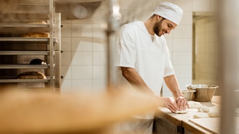 Lächelnde Person in weißem Gewand mit Mütze knetet Teig