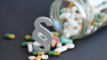 Fokus auf weißes Paragrafenzeichen platziert zwischen verschiedenfarbigen und -förmigen Pillen, die aus vollem umgekippten Rexglas kullern auf grauem Untergrund