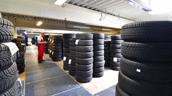 Person in roter Arbeitslatzhose in Autoreifenlager stehend nimmt Notizen auf Clipboard vor, ringsum aufgetürmte Autoreifen