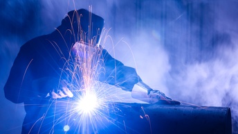 Person mit Schweißhelm und dicken Arbeitshandschuhen beugt sich über ein dickes Metallrohr, um mit einem Schweißgerät dieses zu bearbeiten. Daneben Funken und einen sehr hellen Punkt in der Bildmitte, wo geschweißt wird