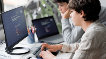 Seitliche Aufnahme zweier Teenager an einem Tisch. Auf dem Tisch steht ein Bildschirm, auf den beide blicken. Daneben sind Kopfhörer, verschiedene Stifte und ein Laptop