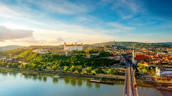 Vogelperspektive einer mehrspurigen Brücke, die über einen Fluss führt. Am Ende der Brücke ist am Ufer eine Stadt mit mehreren Gebäuden. Auf einem Hügel ist eine helle Burg. Am Ufer sind verschiedene Boote