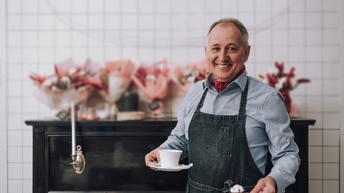 Porträt einer lächelnden Person mit Schürze, die in einer Hand eine Tasse und in der anderen Hand ein kleines Törtchen hält. Hinter der Person stehen auf einem Klavier mehrere Blumensträuße
