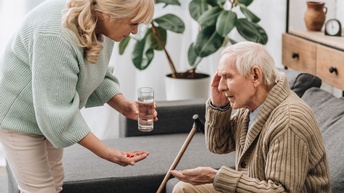 Ältere auf Couch sitzende Person greift sich mit einer Hand an den Kopf und hält andere Hand geöffnet in Richtung vor ihr stehender Person, die sich bückt und in einer Hand Pillen, in anderer Hand Glas mit Wasser hält