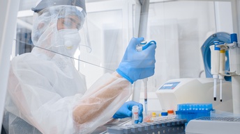 Eine Person sitzt in einem Labor und trägt eine komplette Schutzausrüstung. Sie befüllt mithilfe einer Pipette kleine Proben mit einer Substanz.