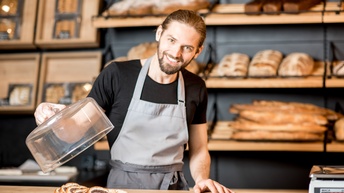 Lächelnde Person mit Schürze steht vor Regal mit Brotlaiben und hebt Deckel über darunterliegendem Kuchen