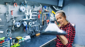 Person in kariertem Hemd hält Schi mit beiden Händen vor sich, kneift eine Auge zu und lugt mit anderem entlang des Schis, im Hintergrund Werkzeugwand mit Werkbank