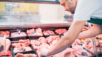 Eine Person mit Handschuhen und Schürze steht neben einer Kühltheke. In der Theke sind verschiedene Fleischstücke in schwarzen Behältern. Auf den Fleischstücken liegen schwarze Schilder. Die Person beugt sich nach vorne und blickt auf das Fleisch