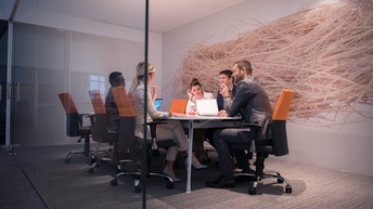 In einem Raum mit einer Glaswand sitzen an einem großen Tisch fünf Personen. Vor manchen stehen Laptops. Die Personen reden miteinander