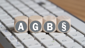 Nahaufnahme des Ausschnitts einer Tastatur, auf der vier Würfel eng nebeneinander liegen. Auf dem ersten ist ein A, auf dem zweiten ein G, auf dem dritten ein B und auf dem vierten ein Paragreifenzeichen