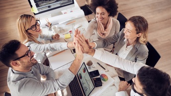 Fünf Personen sitzen um einen Tisch und halten in der Mitte über dem Tisch die Hände zusammen. Auf dem Tisch sind verschiedene Unterlagen, ein Laptop, ein Taschenrechner, Stifte, Handys und Notizblöcke