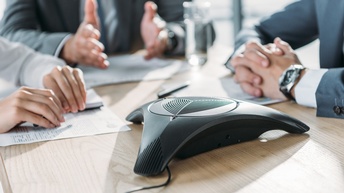 Nahaufnahme eines Telefonlautsprechers auf einem Tisch. Um den Lautsprecher sitzen drei Personen. Vor allen drei Personen liegen Zettel