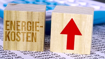 Würfel mit Schriftzug Energiekosten und weiterer Würfel mit Pfeil nach oben stehen auf einem Blatt Papier mit Zahlen
