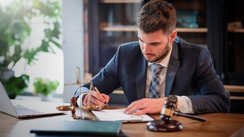 Person in Businesskleidung mit Krawatte und kurzen braunen Haaren sowie Bart sitzt an einem Schreibtisch und unterzeichnet ein Dokument, während daneben ein Richterhammer sowie eine Waage der Gerechtigkeit stehen