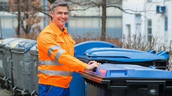 Lächelnde Person in oranger Berufskleidung steht neben Müllcontainern