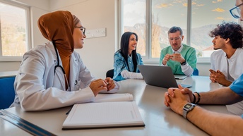 5 Menschen sitzen in einem Raum, um eine Besprechung abzuhalten. Sie blicken einander an und sind in einem Gespräch. Alle tragen Bekleidung und Utensilien, die in Krankenhäusern verwendet werden. 