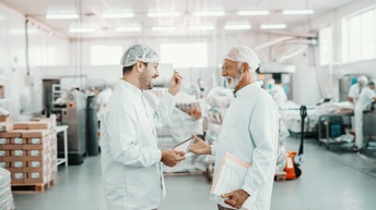 Zwei Personen in weißer Schutzbekleidung mit Haarnetzen unterhalten sich in Lebensmittelproduktionsstätte stehend