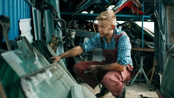 Person in Arbeitslatzhose mit dreckigen Armen in Hockposition greift nach Metallschrott, im Hintergrund Regal mit Fahrzeugteilen