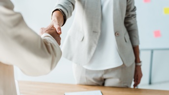 Zwei Personen in hellen Sakkos stehen sich an einem Tisch gegenüber und schütteln sich die Hände. Auf dem Tisch liegt ein Stift auf einem Zettel