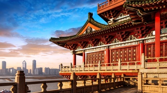 Rechts im Vordergrund ist ein historisches Gebäude mit roten Säulen und spitzen Dächern. Daneben ist ein Gewässer. Am Horizont ist die Skyline der Stadt Peking mit verschiedenen Häusern. Am Himmel sind ein paar Wolken