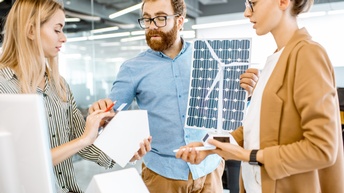 Drei Personen in Gespräch, eine Person hält Modell eines Windrades, ein Person hält Photovoltaikfolie und eine weitere hält Modell eines Hauses, auf dem sie Folie appliziert
