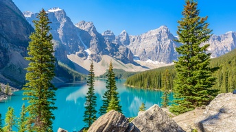 Schroffe Bergkette umsäumt von Nadelwäldern und türkisblauem Gebirgssee, im Vordergrund einzelne Bäume und Felsen